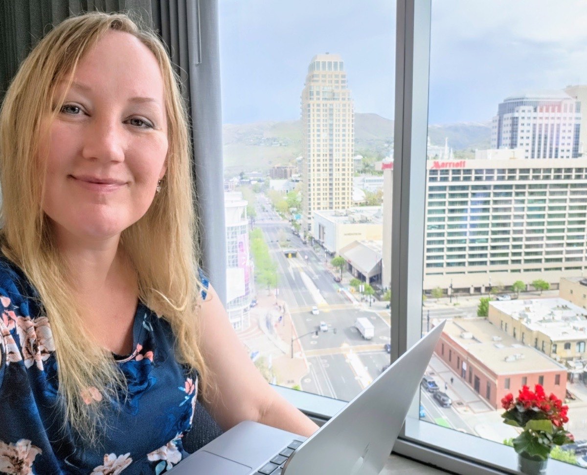 Julie Hurley smiling with her laptop by a window overlooking downtown Salt Lake City